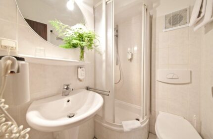 Bathroom at Novum Hotel Leonet Köln Altstadt, a gay-friendly hotel. Sink, shower, toilet, mirror, and fresh flowers. Clean and modern.