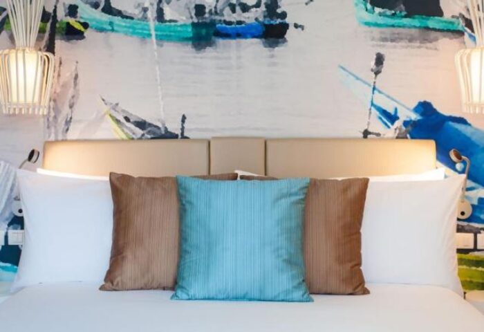 OZO Chaweng Samui gay-friendly hotel room. Bed with pillows, headboard, and wall art featuring boats. Modern lamps.
