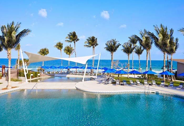OZO Chaweng Samui gay-friendly hotel pool view. Beachfront resort with palm trees, blue umbrellas, and clear blue water. Gay travel.