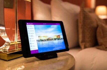 Tablet displaying Okada Manila, a gay-friendly hotel. The screen shows hotel amenities and a view of the Manila skyline. Luxurious room setting.