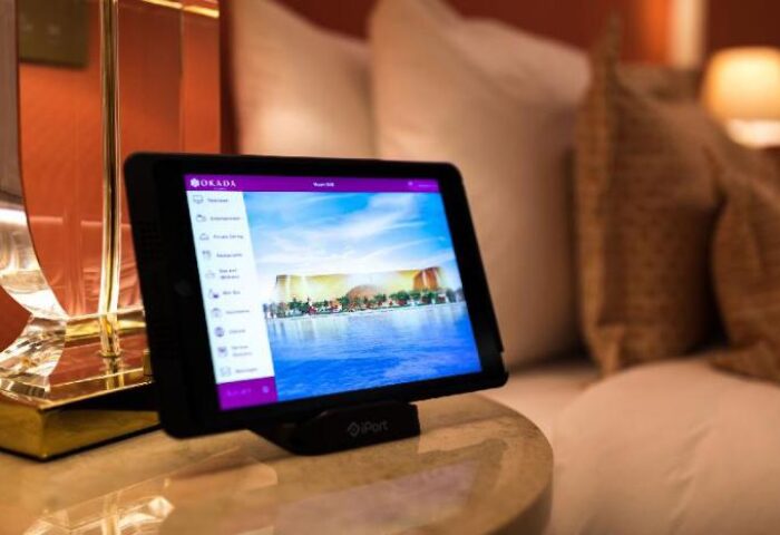 Tablet displaying Okada Manila, a gay-friendly hotel. The screen shows hotel amenities and a view of the Manila skyline. Luxurious room setting.
