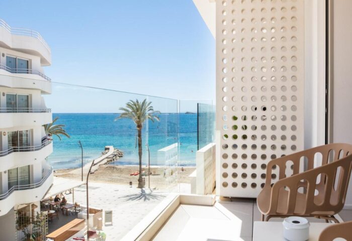 View from a gay-friendly hotel, One Ibiza Suites. Balcony with a chair overlooking the beach and blue sea. Palm trees on the beach.