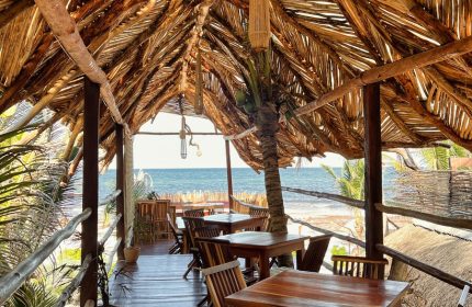 Orchid Beach House Adults Only, gay-friendly hotel. Thatched roof dining area overlooking the ocean. Tables and chairs on a wooden deck.