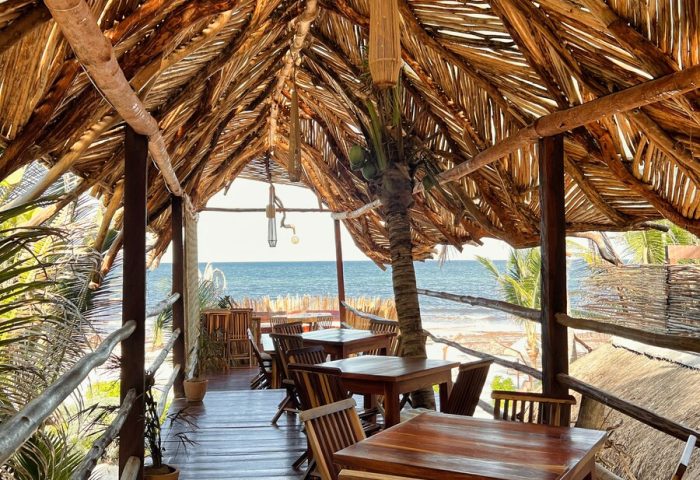 Orchid Beach House Adults Only, gay-friendly hotel. Thatched roof dining area overlooking the ocean. Tables and chairs on a wooden deck.