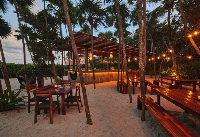Our Habitas Tulum gay-friendly hotel bar area. Tables, chairs, and bar with lights on sandy ground surrounded by palm trees.