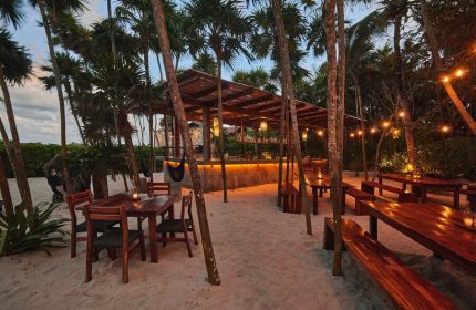 Our Habitas Tulum gay-friendly hotel bar area. Tables, chairs, and bar with lights on sandy ground surrounded by palm trees.