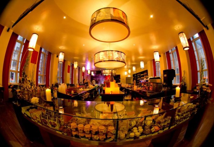 Interior of PLUS Berlin Hotel & Hostel, a gay-friendly hotel. View of the bar area with pastries, warm lighting, and unique hanging lamps.