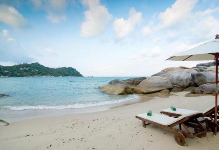 Beachfront view at Panviman Resort Koh Phangan, a gay-friendly hotel. Lounge chairs under an umbrella on the sand. Island view in the distance.