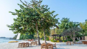 Beachfront dining at Paradee Resort, a gay-friendly hotel. Tables and chairs on the sand under trees offer a relaxing atmosphere.
