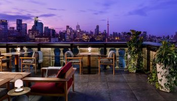 Park Hyatt Toronto's gay-friendly rooftop lounge at dusk. City skyline view, tables, chairs, and plants create an inviting atmosphere.