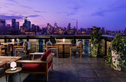 Park Hyatt Toronto's gay-friendly rooftop lounge at dusk. City skyline view, tables, chairs, and plants create an inviting atmosphere.