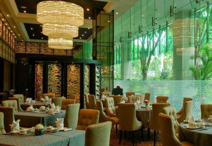 Elegant dining room at Park Regis Singapore, a gay-friendly hotel. Tables are set beneath a grand chandelier. Lush greenery visible.