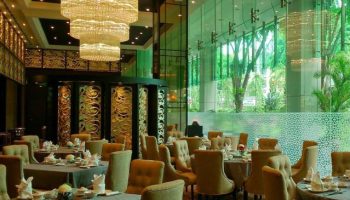 Elegant dining room at Park Regis Singapore, a gay-friendly hotel. Tables are set beneath a grand chandelier. Lush greenery visible.