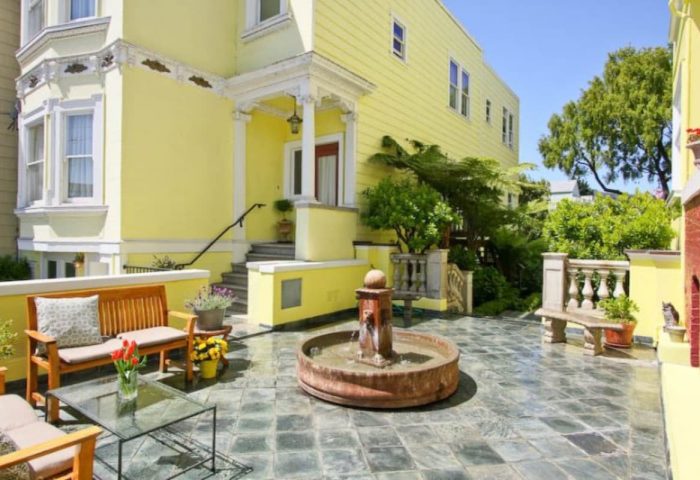 Parker Guest House's courtyard, a gay-friendly hotel. Yellow building with a fountain, benches, and lush greenery. A serene retreat for gay men.