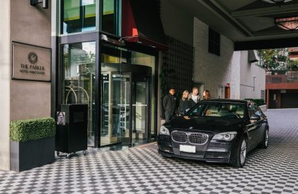 Exterior of the gay-friendly Parker Hotel Vancouver, showcasing the entrance with a revolving door, a black car, and guests arriving.