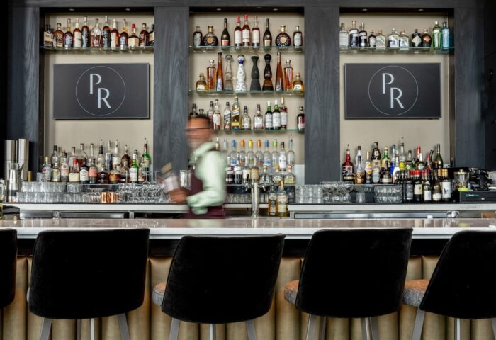 Bar area at the gay-friendly Parker Hotel Vancouver. Shelves display bottles. Bartender in motion. Black bar stools in the foreground.