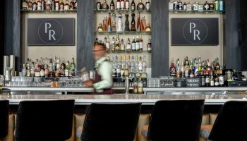 Bar area at the gay-friendly Parker Hotel Vancouver. Shelves display bottles. Bartender in motion. Black bar stools in the foreground.