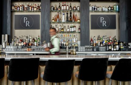 Bar area at the gay-friendly Parker Hotel Vancouver. Shelves display bottles. Bartender in motion. Black bar stools in the foreground.