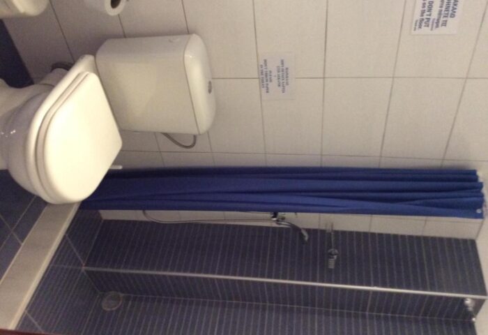 Bathroom at Pension Ageliki Kalogera, a gay-friendly hotel. Toilet, shower with blue curtain, and tiled walls. Clean and simple design.