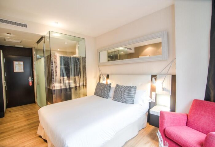 Petit Palace Chueca gay-friendly hotel room. Modern design with glass-enclosed shower, double bed, pink chair, and stylish decor. Madrid lodging.