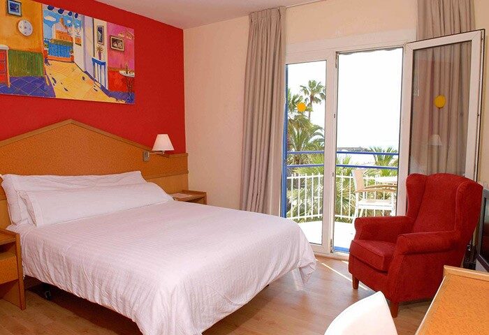 Gay-Friendly Hotel Platjador room. Balcony view, red accent wall, and comfy armchair. Ideal for gay travelers seeking comfort and style.