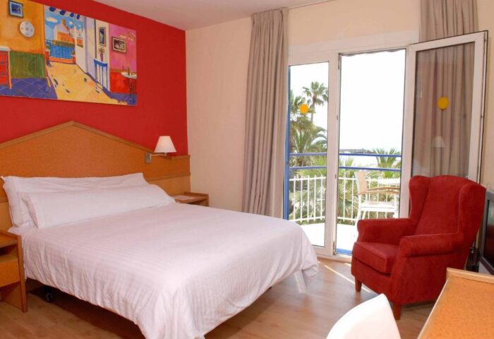 Platjador gay-friendly hotel room with balcony, bed, and red armchair. A colorful painting hangs on the red wall. Palm trees visible outside.