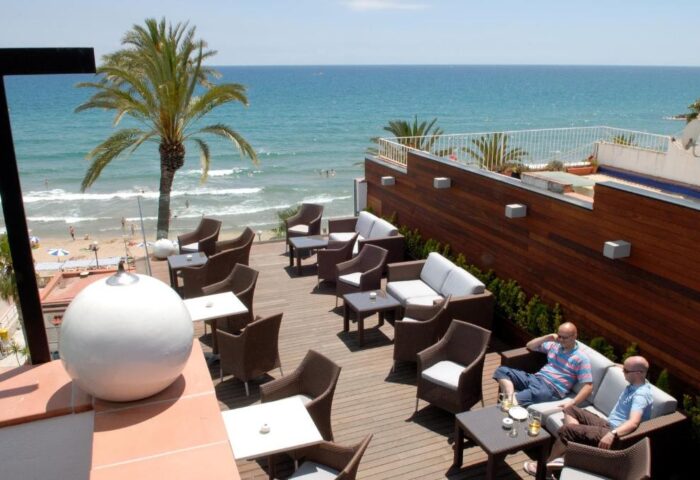 Gay-Friendly Hotel Platjador rooftop lounge area with ocean view. Men relax on sofas, enjoying the beach scenery. Ideal gay travel destination.