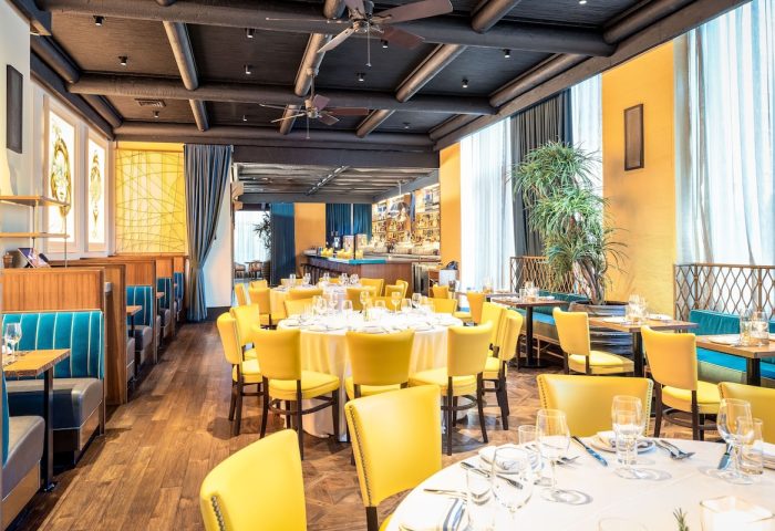 Pod Times Square gay-friendly hotel restaurant. Yellow chairs surround tables with white linens. Booth seating lines the left wall. Bar in background.