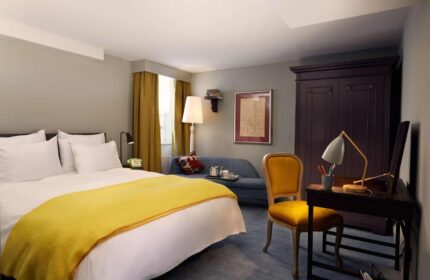 Pulitzer Amsterdam gay-friendly hotel room. Features a bed with yellow blanket, desk, chair, sofa, and wardrobe. Gay travel advice.
