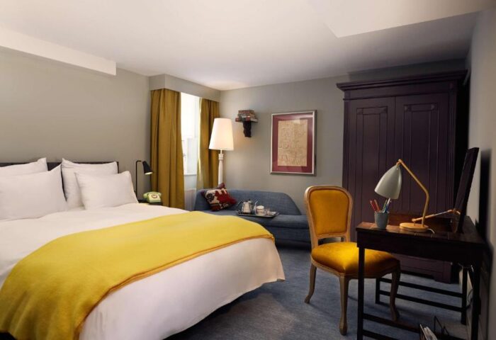 Pulitzer Amsterdam gay-friendly hotel room. Features a bed with yellow blanket, desk, chair, sofa, and wardrobe. Gay travel advice.