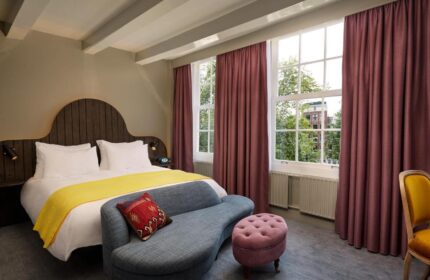 Pulitzer Amsterdam gay-friendly hotel room. Bed with yellow throw, blue sofa, and pink ottoman. Windows with maroon curtains. Stylish decor.