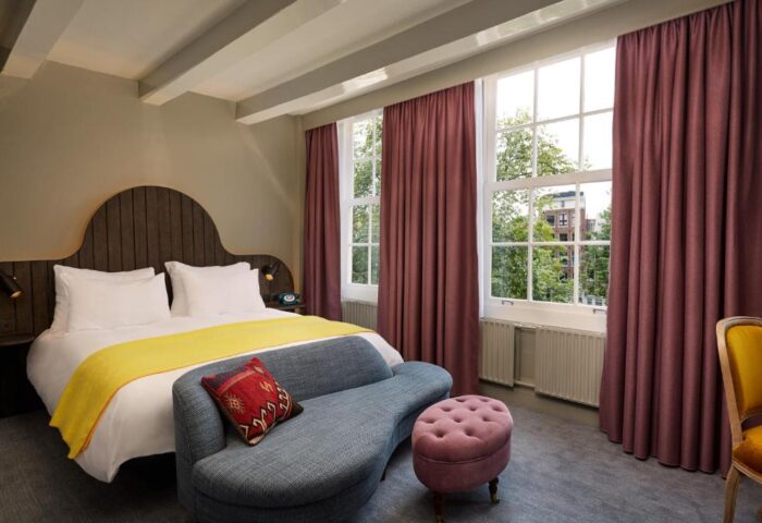 Pulitzer Amsterdam gay-friendly hotel room. Bed with yellow throw, blue sofa, and pink ottoman. Windows with maroon curtains. Stylish decor.