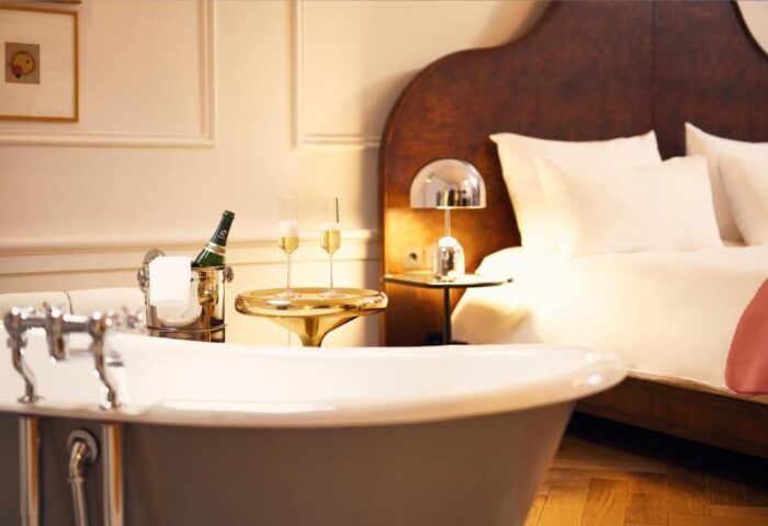 Pulitzer Amsterdam gay-friendly hotel room. Bathtub, champagne bucket, and flutes on a gold table. Bed with white linens in background.