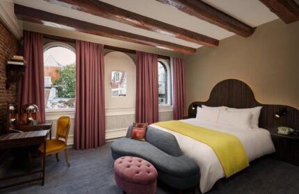 Gay-friendly Pulitzer Amsterdam hotel room. Beams, arched windows with curtains, desk, chair, bed with yellow throw, and a blue couch.