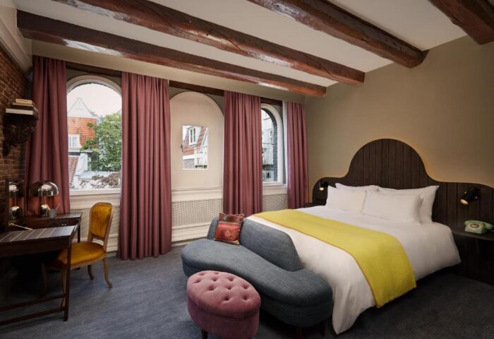 Gay-friendly Pulitzer Amsterdam hotel room. Beams, arched windows with curtains, desk, chair, bed with yellow throw, and a blue couch.