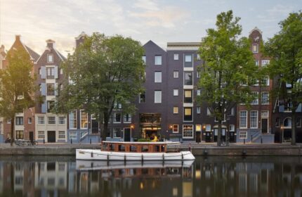 Pulitzer Amsterdam, a gay-friendly hotel. Canal view with a boat. Amsterdam's architecture reflects in the water. Trees add to the charm.