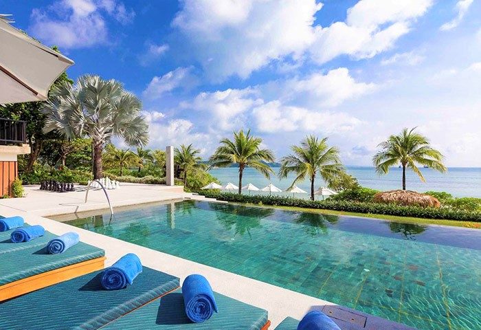 Pullman Phuket Panwa Beach Resort pool view. Gay-friendly hotel with SHA certification. Palm trees, turquoise water, and blue towels.