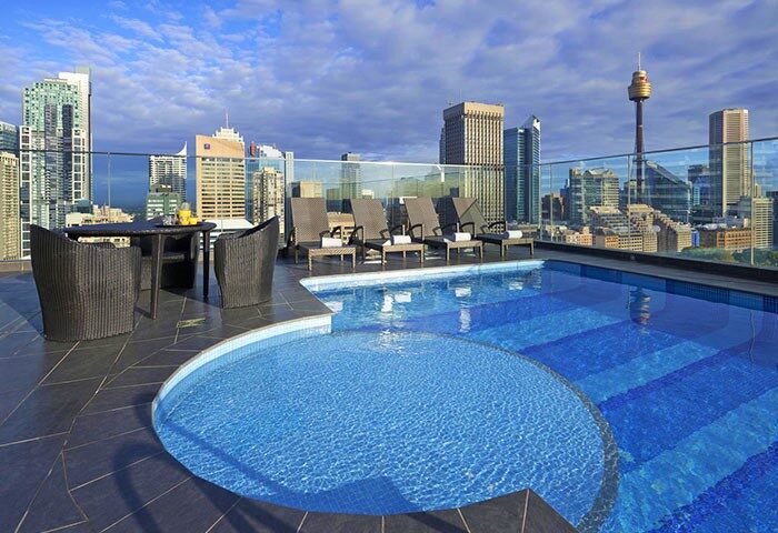 Pullman Sydney Hyde Park gay-friendly hotel rooftop pool. Sydney skyline view from hotel pool deck with lounge chairs and table seating.