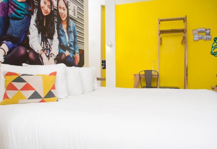 Qbic London City gay-friendly hotel room. Bright yellow wall, bed with geometric pillow, and a mural of three young women. Pet-friendly.