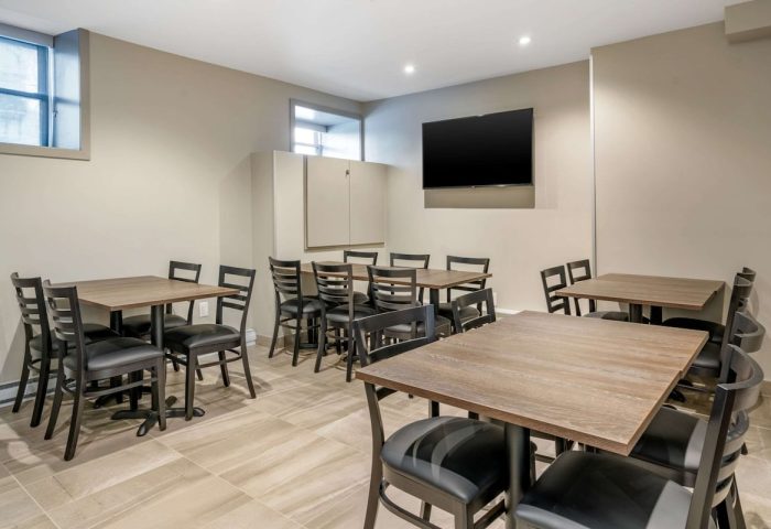 Quality Inn Centre-Ville gay-friendly hotel dining area. Tables with black chairs, a TV, and storage in a neutral-toned room.