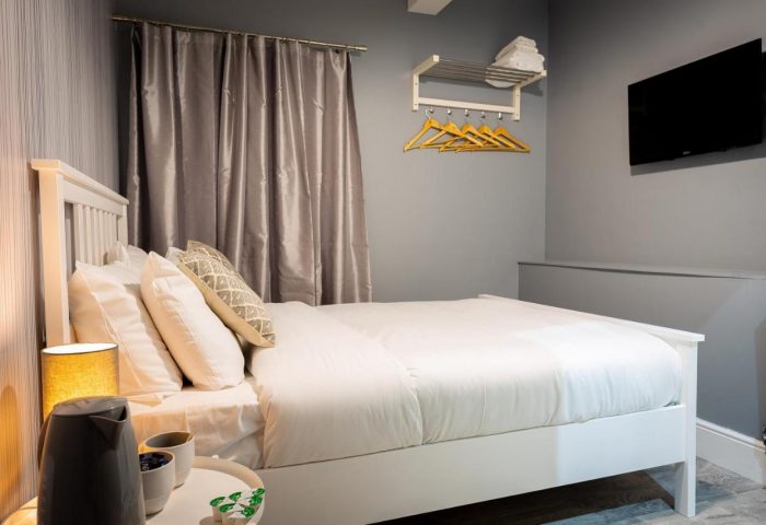 REM Hotel bedroom. Gay-friendly hotel room with a white bed, gray walls, TV, hangers, and bedside table with kettle. Cozy, modern design.