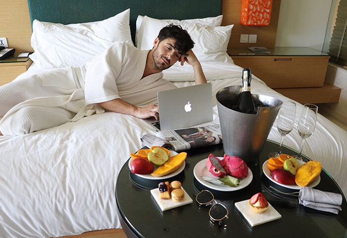 Gay man in robe using laptop in bed at Renaissance Phuket Resort & Spa. Fruit, pastries, champagne, and sunglasses on table. Luxury travel.