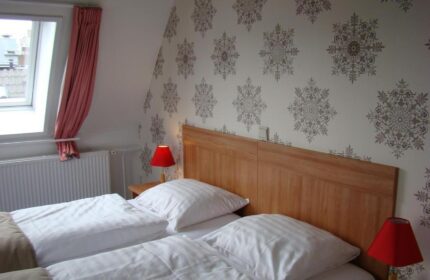 Rho Hotel gay-friendly room. Twin beds with white linens, wood headboard, patterned wallpaper. Red lamps on nightstands. Cozy gay hotel room.