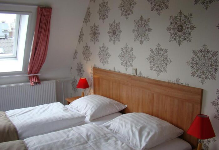 Rho Hotel gay-friendly room. Twin beds with white linens, wood headboard, patterned wallpaper. Red lamps on nightstands. Cozy gay hotel room.