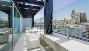Room Mate Oscar gay-friendly hotel balcony view. Madrid skyline visible. White chairs, glass railing, modern design. Gay travel accommodation.