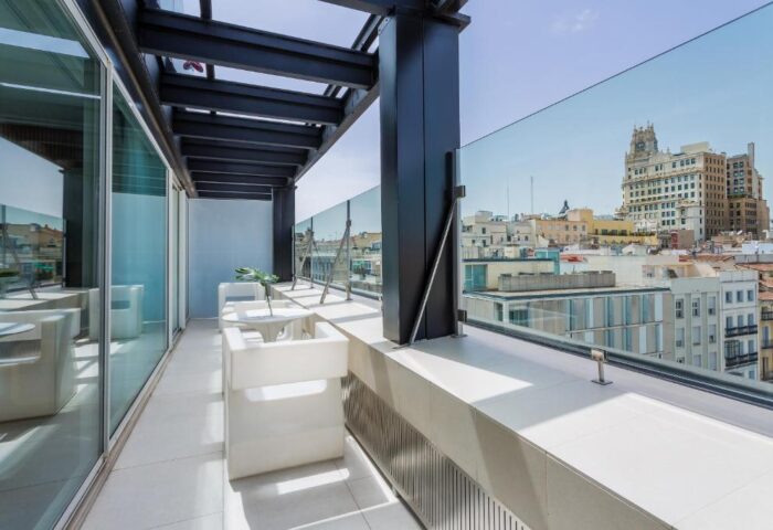 Room Mate Oscar gay-friendly hotel balcony view. Madrid skyline visible. White chairs, glass railing, modern design. Gay travel accommodation.