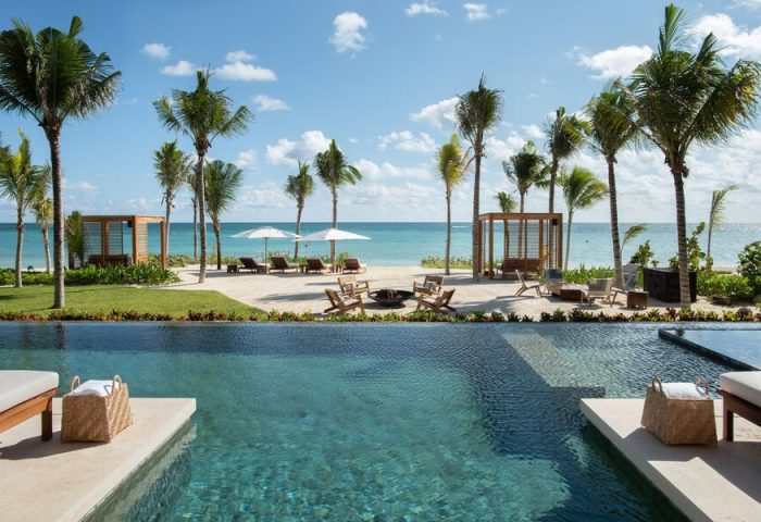 Rosewood Mayakoba gay-friendly hotel view. Poolside lounge area with palm trees, beach, and ocean. Luxury travel destination for gay men.