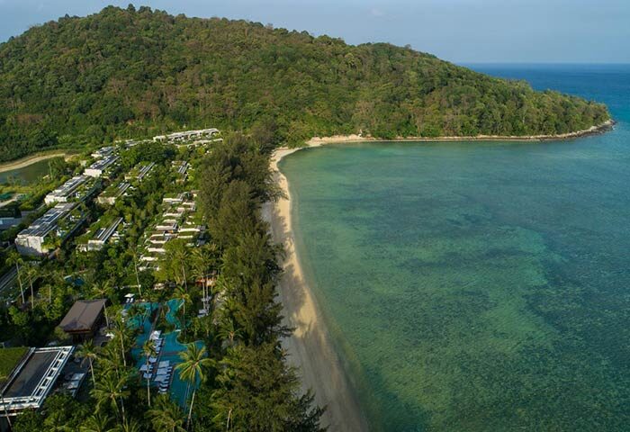 Aerial view of Rosewood Phuket, a gay-friendly hotel. Lush greenery surrounds the resort, nestled along a pristine beach with clear turquoise waters.