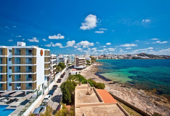 Ryans Ibiza Apartments, gay-friendly, adults-only hotel. Coastal view with blue sky, turquoise sea, and modern hotel architecture.