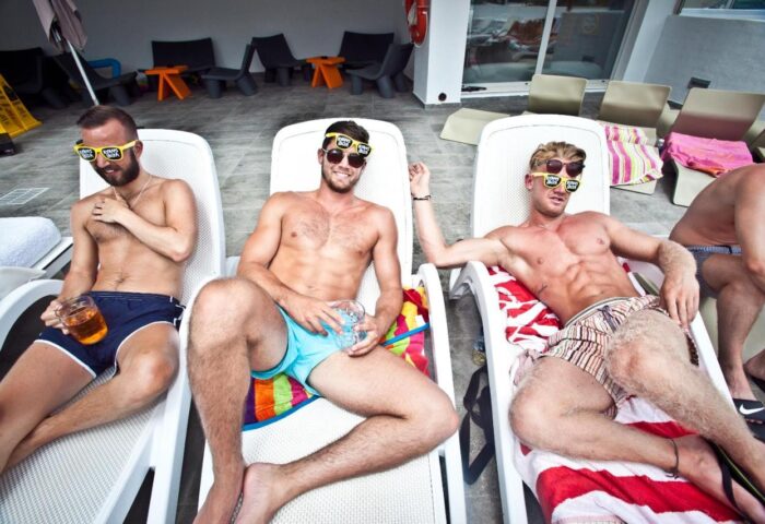Gay-Friendly Ryans Ibiza Apartments: Adults relax by the pool. Men in swimwear and sunglasses enjoy drinks at this Ibiza hotel.